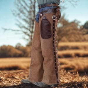 May include: Tan suede leather chaps featuring a dark brown leather pocket on the thigh, a silver metal ring at the hip, and long fringe detailing along the outer leg seams. The chaps are secured with a series of metal studs down the side.