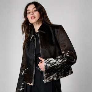 May include: A jacket featuring a mix of dark brown and black cowhide textures with white mottled patterns on the sleeves and lower panels. The garment has a classic collar, a full-length front zipper, and long sleeves, worn over a black shirt and pants.
