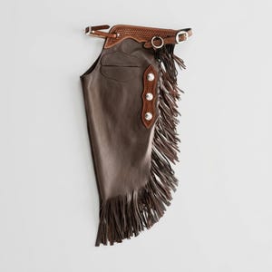 May include: A dark brown leather chap featuring a tan leather yoke with basket-weave tooling, a silver-toned metal buckle, and a D-ring. The side is adorned with a vertical leather strip containing three silver-toned conchos and long fringe.