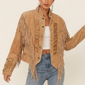Peut inclure: Une veste en daim beige avec de longues franges sur la poitrine et les manches. La veste présente un col montant, une fermeture boutonnée sur le devant et des détails de laçage décoratifs sur les panneaux avant et les poignets, portée sur un haut blanc.