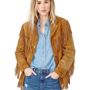 May include: A tan suede jacket featuring long fringe detailing along the chest, shoulders, and sleeves. The jacket has a collared neckline, a button-front closure, and long sleeves. It is worn over a light blue button-down shirt and blue jeans.