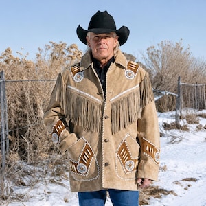May include: A tan suede jacket featuring long fringe across the chest and shoulders, accented with brown leather patches, white stitching, and circular beaded embellishments on the front pockets and upper chest, worn with a black cowboy hat.