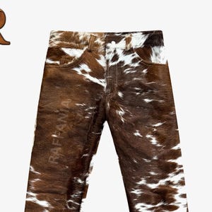 Puede incluir: Pantalones de piel de vaca con un estampado de manchas marrones y blancas. Los pantalones tienen un diseño clásico de cinco bolsillos con cierre de botón y cremallera. La palabra "RAFFANIA" está impresa en la pierna izquierda.