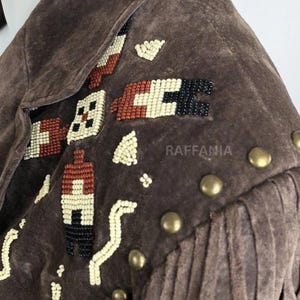 May include: A brown suede jacket featuring intricate beadwork in cream, rust, and black geometric patterns on the shoulder. The garment includes a row of round brass-toned metal studs along the shoulder seam and long suede fringe detailing.