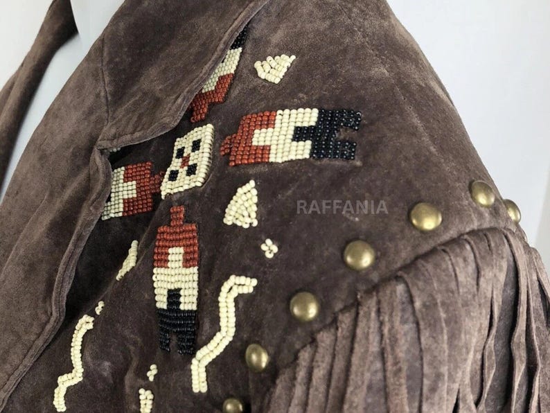 May include: A brown suede jacket featuring intricate beadwork in cream, rust, and black geometric patterns on the shoulder. The garment includes a row of round brass-toned metal studs along the shoulder seam and long suede fringe detailing.