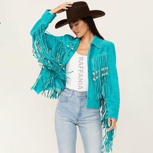 Puede incluir: Una chaqueta de ante color turquesa brillante con largos flecos en el pecho y las mangas, adornada con peque&ntilde;as cuentas cil&iacute;ndricas. La chaqueta se lleva sobre una camiseta blanca con el texto RAFFANIA impreso verticalmente en gris claro.