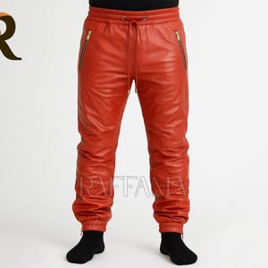 May include: Red leather joggers with gold zippers on the pockets and ankles. The pants have a drawstring waist and are worn with black socks. The pants are made of a shiny leather material.