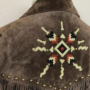 May include: A brown suede jacket featuring a beaded geometric star pattern on the back. The design includes cream, black, and rust-colored beads. A row of brass-toned metal studs follows the yoke seam, and long fringe hangs from the bottom edge.