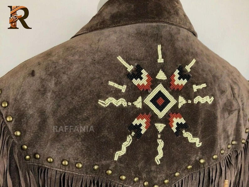 May include: A brown suede jacket featuring a beaded geometric star pattern on the back. The design includes cream, black, and rust-colored beads. A row of brass-toned metal studs follows the yoke seam, and long fringe hangs from the bottom edge.