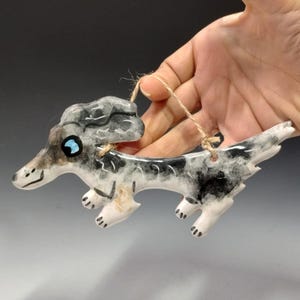 May include: A ceramic ornament of a grey and white dachshund with blue eyes. The dachshund is running and has a string attached to its back for hanging.