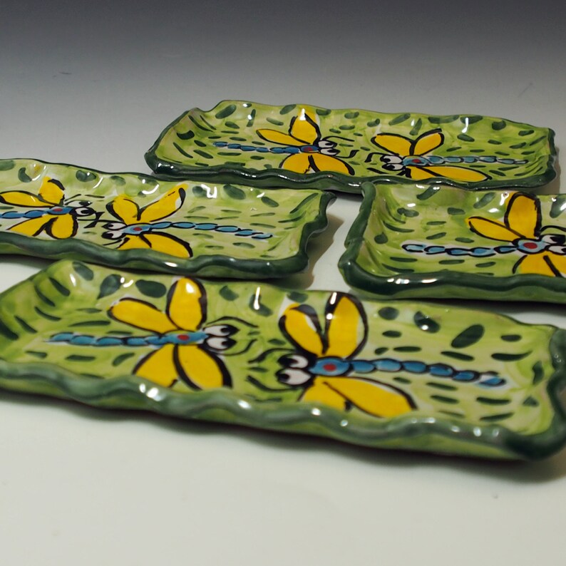 May include: Four rectangular ceramic trays with wavy edges, each featuring a hand-painted green background with dark green brushstrokes and a central illustration of a yellow dragonfly with a blue body and large white eyes with black pupils.