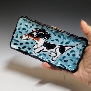 May include: Hand-painted ceramic dachshund dog tray. The rectangular tray features a light blue background with dark blue polka dots. The dachshund is black, white, and brown. The tray has a black border.