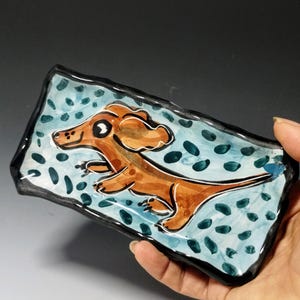 May include: A ceramic dish with a blue and white background and a brown dachshund painted on it. The dachshund has a big eye and is running.