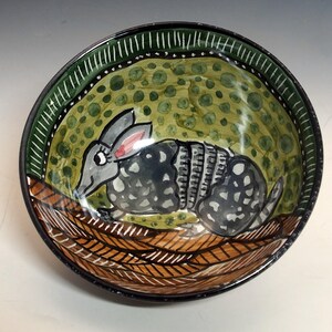 May include: Hand-painted ceramic bowl featuring an armadillo design. The bowl has a black rim with a green and white patterned border. The interior has a green background with dark green spots and a brown and white patterned base.