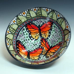 May include: Hand-painted ceramic bowl featuring three orange and black monarch butterflies against a light green dotted background. The rim has a decorative leaf pattern in green, black, and orange. The bowl is round and has a black rim.