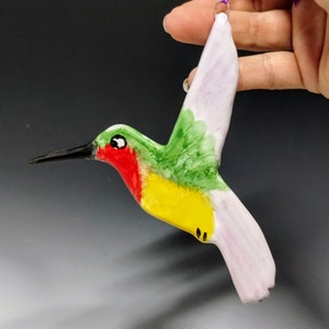 May include: A hand-painted ceramic hummingbird ornament with green, yellow, red, and white paint. The hummingbird is in flight with its wings spread out.