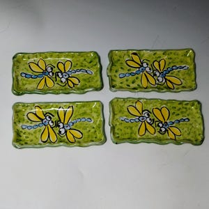 May include: Four rectangular ceramic trays with wavy edges, each featuring a hand-painted design of two yellow dragonflies with blue bodies against a mottled light green background. The trays are arranged in a two-by-two grid pattern.