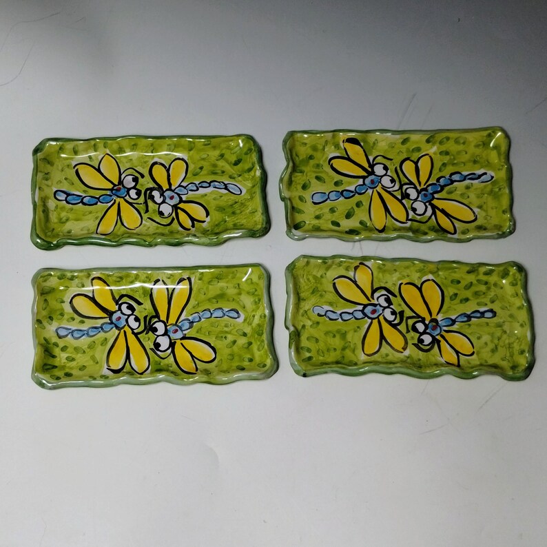 May include: Four rectangular ceramic trays with wavy edges, each featuring a hand-painted design of two yellow dragonflies with blue bodies against a mottled light green background. The trays are arranged in a two-by-two grid pattern.