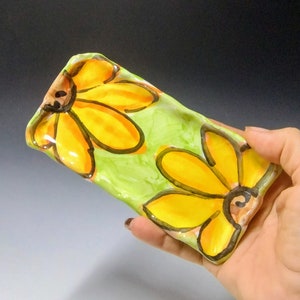 May include: A small, rectangular ceramic dish with a green background and two yellow flowers with black outlines. The dish is glazed and has a slightly textured surface.