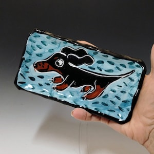 May include: Rectangular ceramic dish with a black and brown dachshund dog illustration on a light blue background with dark blue accents. The dish has a black border and is held by a hand.