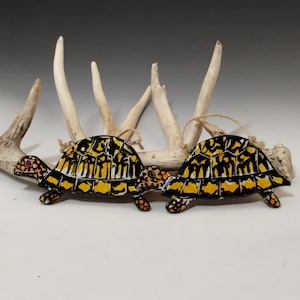 May include: Two ceramic turtle ornaments with black, yellow, and white painted details. The turtles are hanging from a white antler.