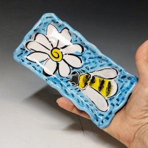 May include: Hand-painted ceramic dish with a blue background and a white daisy and a bee design. The rectangular dish has a wavy edge. The bee has yellow and black stripes. The daisy has a yellow center.