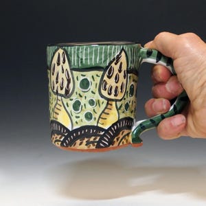 May include: A ceramic mug with a hand-painted mushroom design. The mug has a green and white striped rim, a green handle with black stripes, and a base with brown and white details. The mushroom design includes yellow, brown, and green colours.