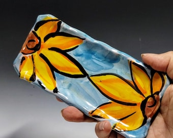 May include: A handmade ceramic dish with a blue glaze and two yellow sunflowers painted on it.