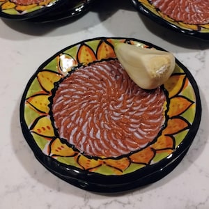 May include: A set of ceramic plates with a sunflower design. Each plate has a black rim, a yellow and orange floral pattern, and a textured center. One plate holds a garlic clove.