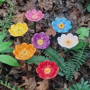 Puede incluir: Una colección de siete estacas de jardín con forma de flor de cerámica colorida. Las flores son en tonos rosa, amarillo, azul, morado, naranja, blanco y rojo, cada una con un centro amarillo. Las estacas están colocadas entre follaje verde y hojas marrones.
