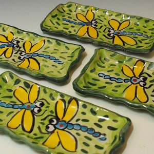May include: Four rectangular ceramic trays with wavy edges, each featuring a hand-painted light green background with dark green dash patterns and a central illustration of a blue-bodied dragonfly with large yellow wings and black eyes.