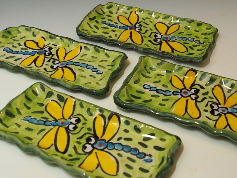 May include: Four rectangular ceramic trays with wavy edges, each featuring a hand-painted light green background with dark green dash patterns and a central illustration of a blue-bodied dragonfly with large yellow wings and black eyes.