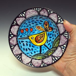 May include: A hand-painted ceramic plate featuring a yellow bird with orange and black details against a blue background with black dots. The plate has a black rim with a decorative purple and white floral pattern. The plate is held by a hand.