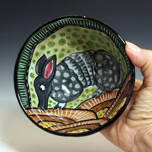 May include: Hand-painted ceramic bowl featuring an armadillo design. The bowl has a black rim with a green and white border. The interior has a green background with brown and yellow accents. The armadillo is gray with black spots.