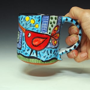 K&ouml;nnte beinhalten: Ein Set aus drei Keramikbechern mit einer gesprenkelten, cremefarbenen Glasur. Jeder Becher hat ein anderes handgemaltes Design: ein blaues und gr&uuml;nes Blatt, eine gelbe Blume und eine rote Beere. Die Becher haben ein schlichtes, modernes Design.