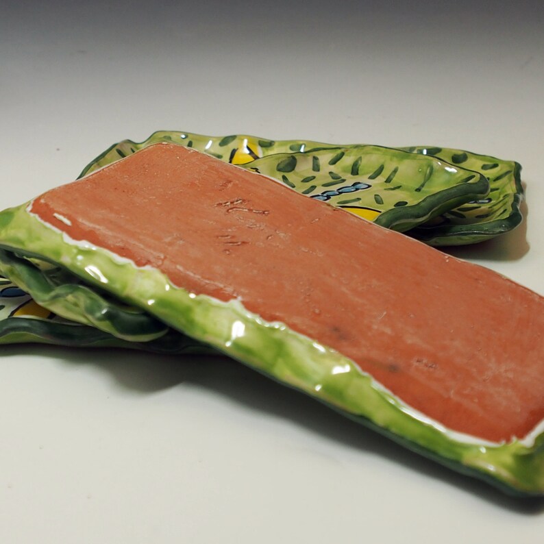 May include: A rectangular ceramic tray with a terracotta underside and a hand-painted green, yellow, and blue patterned top surface. The edges are wavy and glazed in a glossy green finish, with a signature etched into the clay base.