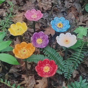 May include: A collection of colorful ceramic flower garden stakes in shades of pink, yellow, blue, purple, orange, white, and red. Each flower has a detailed center and is set against a backdrop of leaves and foliage.