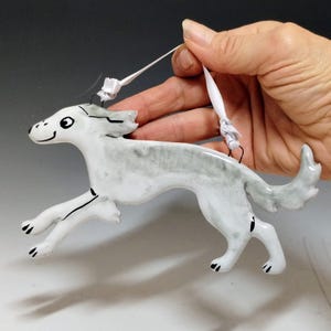 May include: A ceramic dog ornament in a running pose, painted in shades of white and gray with black accents. The dog has a smiling expression and is suspended by a white ribbon. A decorative item for pet lovers.
