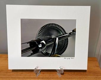 May include: Close-up of a vintage chrome hand mixer, a kitchen tool for mixing ingredients. The mixer is mounted in a white mat, displayed on a wooden surface. The image is a photograph.
