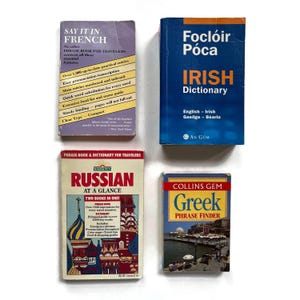 May include: Four vintage language books are displayed on a white surface. The books include a French phrase book, an Irish dictionary, a Russian phrase book, and a Greek phrase finder. The books are compact and feature colorful covers.
