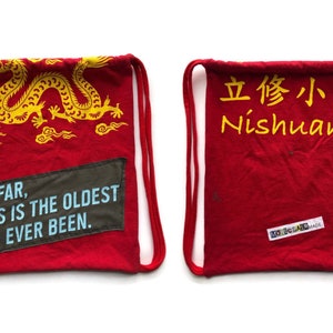 May include: Red drawstring bag with a yellow dragon design and the text "SO FAR, THIS IS THE OLDEST I'VE EVER BEEN." on one side and the text "立修小學 Nishuane" on the other side.