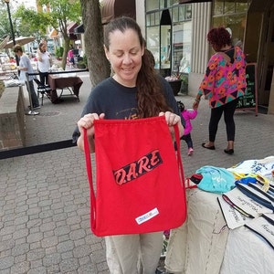Könnte beinhalten: Eine rote Kordeltasche mit dem Wort "DARE" in schwarzen Buchstaben bedruckt. Die Tasche wird von einer Person gehalten.