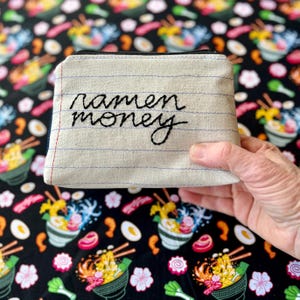 May include: A beige canvas coin purse with the embroidered words "ramen money" in black script. The purse has a zipper closure and is lined with blue lines. The background is a black fabric with a ramen bowl pattern.