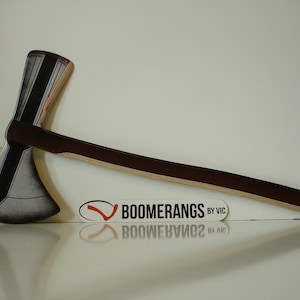 May include: A wooden throwing axe with a black and grey painted blade and a brown wooden handle. The axe is on a white surface with a logo that says "Boomerangs by Vic".