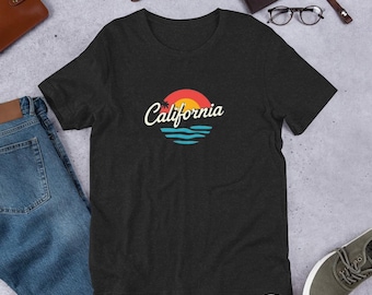 May include: Dark gray t-shirt featuring a sunset design with the word "California" in a script font. The graphic includes a red and orange sun, blue waves, and a palm tree silhouette. The shirt is displayed on a flat surface with other accessories.