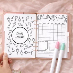 May include: A pink planner open to a July calendar page with a doodle theme. The left page has a "Daily Doodle" circle surrounded by marshmallow illustrations. The right page includes a calendar, notes section, and two highlighters.
