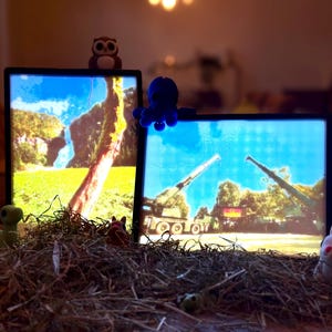 Op de afbeelding: Twee ingelijste foto's met verschillende scènes. De foto links toont een groen veld met een grote boom en een blauwe lucht. De foto rechts toont een militair voertuig met een groot kanon. Beide foto's zijn van achteren verlicht.