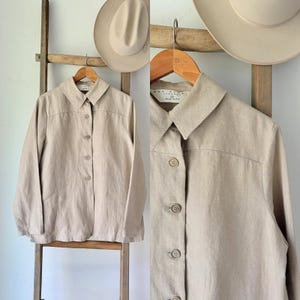 May include: A beige button-down shirt with a collar, hanging on a wooden hanger. The shirt has a classic design with buttons down the front and is made of a light-colored fabric. Two beige hats are hanging in the background.