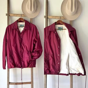 May include: A burgundy nylon jacket with a white interior lining, displayed on a wooden hanger. The jacket has a button-up front, pockets, and a drawstring at the bottom. Two hats hang above the jacket on a wooden ladder.