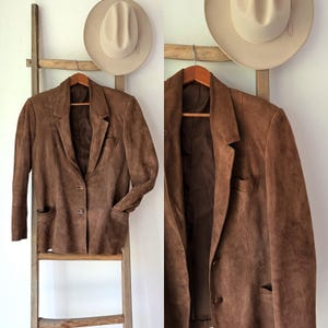 Op de afbeelding: Een bruine suède blazer met een sluiting met twee knopen en twee voorzakken hangt aan een houten hanger. Een lichtgekleurde hoed hangt boven de blazer. De blazer wordt op een houten ladder getoond.